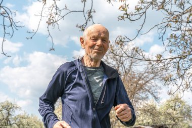 90 yaşında spor giyimli bir adam bahar güneşli bir günde çiçek açan bahçede koşuyor.