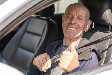 Kablosuz kulaklıklı yaşlı mutlu adam araba kullanıyor ve baş parmağını kaldırıyor.