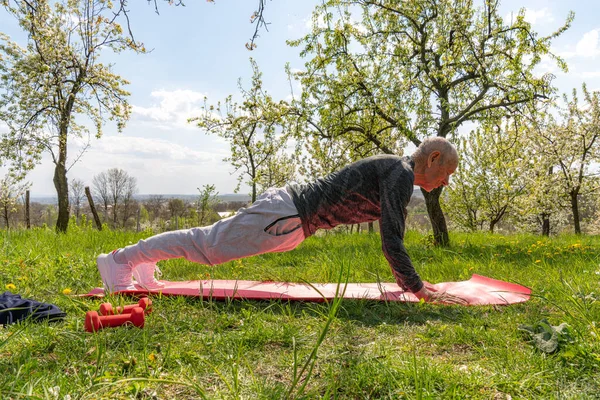 Full body of old sportsman doing plank on stadium. Profile of man in training suit and white sneakers standing in plank position.