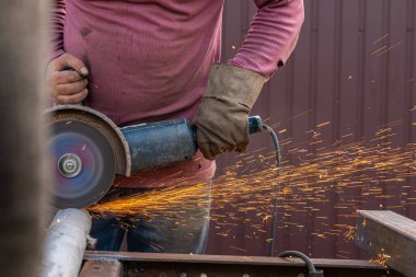 Yanlarda kaynak makinesinden parlak kıvılcımlar uçuşuyor. Çalışan eldiven giymiş genç bir kaynakçı garajda metal bir ürün öğütüyor.