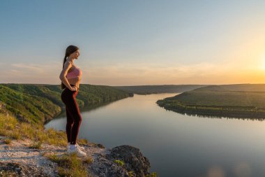 Spor giyim ve gözlüklü genç aktif kadın yüksek irtifada bir kayanın üzerinde dinleniyor, gün batımlı güzel bir manzara izliyor.