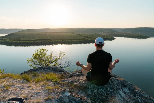 Yogada kaya başında oturan genç adam ruhani rahatlama için poz veriyor dinginlik ve nefes kesici nehirde meditasyon yapıyor. — Stok fotoğraf