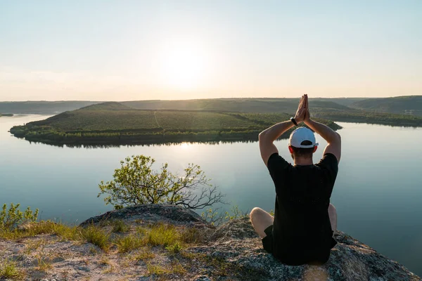 Erkek yürüyüşçü, geniş nehrin yanındaki kayada yoga yapıyor. Bir adam dağda Tanrı 'ya dua ediyor. — Stok fotoğraf