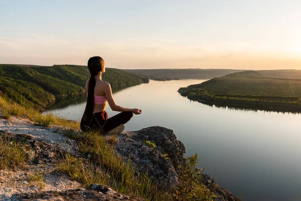 Брюнетка практикует йогу и сидит в позе лотоса на открытом воздухе с удивительным видом на реку . — стоковое фото