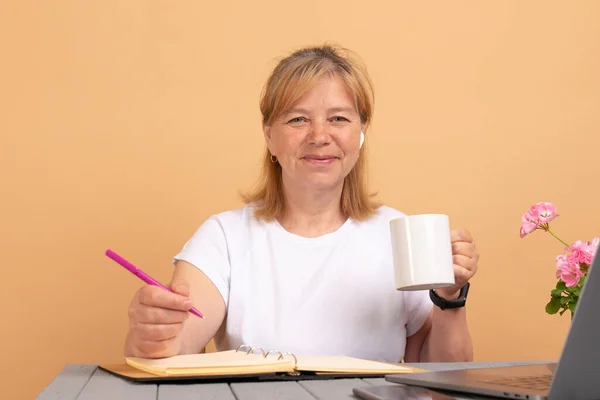 Mulher otimista em fones de ouvido sem fio e roupas brancas casuais bebendo xícara de café fresco enquanto sentado na mesa de madeira perto do laptop e olhando para a câmera — Fotografia de Stock