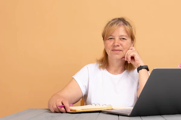 Atraente positivo talentoso mulher madura escritor sentado na frente do laptop e escrever seu novo livro, olhando para a câmera com sorriso alegre — Fotografia de Stock