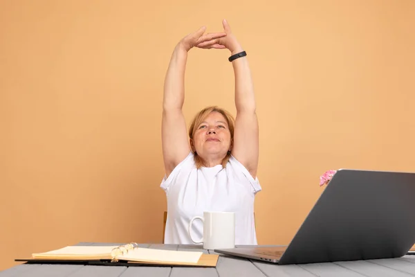 Senior woman sitting at the table with laptop, resting with raised hands, showing hairy unshaven female armpits. Body positive trend