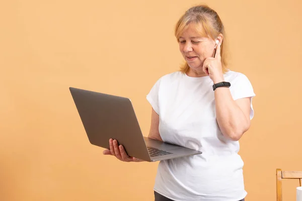 Avó na era Digital. Close-up de mulher sênior pensativo tentando lidar com o computador, enquanto em pé contra o fundo laranja — Fotografia de Stock