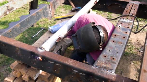 A strong man is a welder in a red T-shirt, in a welding mask and welders leathers, a metal product is welded with a welding machine in the garage — Stock Video