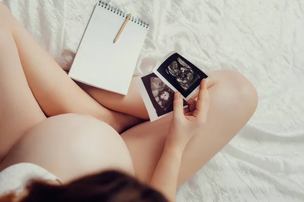 Uma bela mulher grávida senta-se em sua cama e olha para o pict — Fotografia de Stock