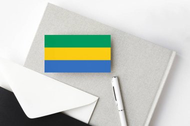 Minimalist arka planda Gabon bayrağı. Beyaz kalemli ve defterli ulusal davetiye zarfı. İletişim konsepti.