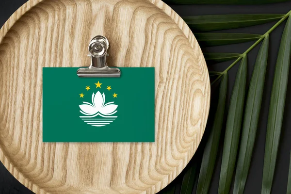 Macao flag tagged on wooden plate. Tropical palm leaves monstera on background. Minimal national concept.