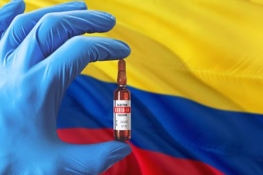 Coronavirus Covid-19 konseptli Kolombiya bayrağı. Mavi koruma eldivenli doktor elinde aşı şişesi tutuyor. Salgın virüsü, Cov-19, Corona virüsü salgını.
