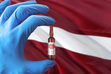 Coronavirus Covid-19 konseptli Letonya bayrağı. Mavi koruma eldivenli doktor elinde aşı şişesi tutuyor. Salgın virüsü, Cov-19, Corona virüsü salgını.