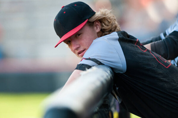 High school baseball player with long hair leaning on dugout fen