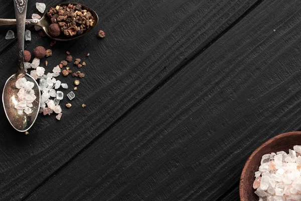 Spoons with Himalayan salt and seasonings on a black wooden background and a saucer with salt. With place for inscription