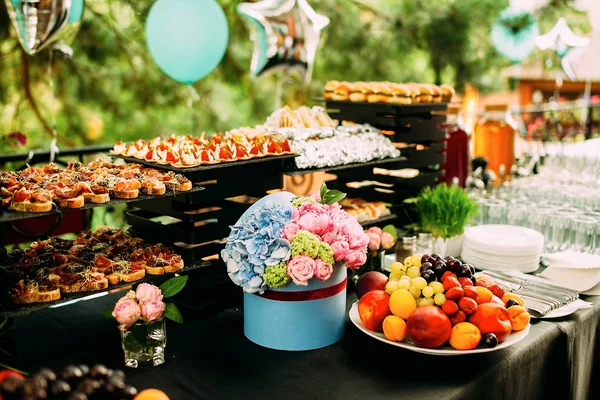 A table with a variety of snacks and slices. Food at an open-air party. Mini burgers and canapes. Decorated table with food flowers and grass. — Stock Photo, Image