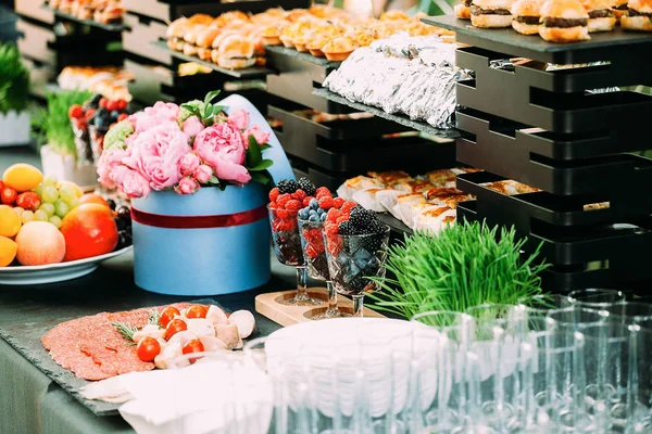 Un tavolo con una varietà di snack e fette. Cibo a una festa all'aperto. Mini hamburger e tartine. Tavolo decorato con fiori alimentari ed erba . — Foto Stock