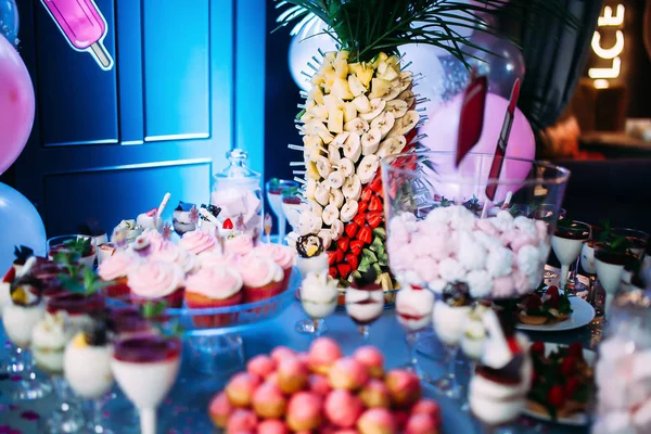 Schön dekorierter Tisch mit verschiedenen Kuchen und Süßigkeiten. Eis mit Marmelade und Minze in Gläsern. Kanapees mit Früchten. ein großer Teller mit rosa bis. Glas mit rosa Marshmallows. Palme mit Früchten. — Stockfoto