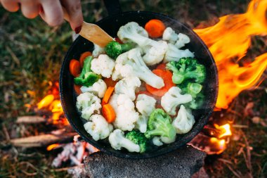 Karnabahar, brokoli ve tavada havuç. Açık ateşte yemek pişirmek. Açık hava yemekleri. Izgara sebze. Kamp gezisinde yemek. Spatulalı bir el. Fotoğraf üstten.