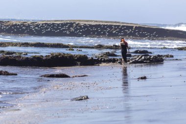 Siyah elbiseli ve barakalı genç bir kadın ıslak sahilde yürüyor Şili sahillerinde doğal manzaranın içinde, Pupuya, Navidad, O 'Higgins, Şili