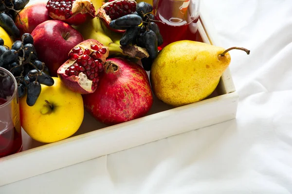 बेडशीटवर पांढर्या लाकडी ट्रेमध्ये सफरचंद, मोती, केळी, द्राक्षे आणि डाळिंबाच्या फळांसह लाल ताजे रस. शीर्ष दृश्य. निरोगी जीवनशैली संकल्पना — स्टॉक फोटो, इमेज