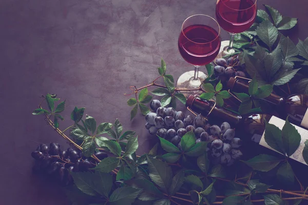Натюрморт с бокалами, бутылками, виноградом и листьями на коричневом деревянном столе — стоковое фото