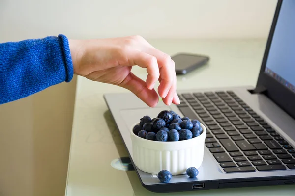 Young woman using laptop computer at home and eating blueberry.