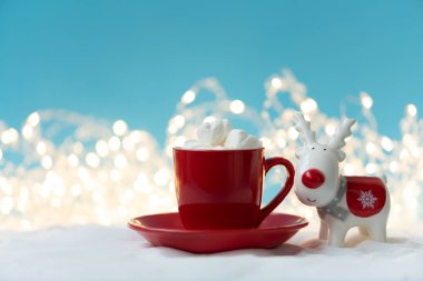 Kırmızı bardakta kremalı sıcak çikolata. Karlı arka planda noel süsleri çıkarılmış ren geyiği oyuncağı. Noel ve yeni yıl tatili konsepti. Boşluğu kopyala