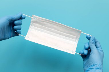 Mavi lateks eldiven giyen ve mavi arka planda cerrahi kulak ilmiği maskesi takan doktorların elleri. Koruma kavramı