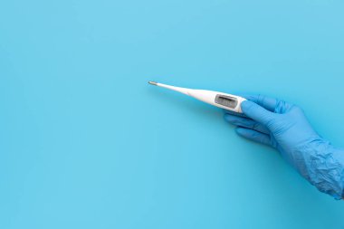 Mavi lateks eldiven takan ve elinde elektrikli termometre tutan sıhhiyeci el mavi arka planda düşük sıcaklık gösteriyor. Grip, hastalık konsepti