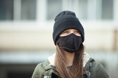 Kulak ilmeği maskeli ve siyah şapkalı genç bir kızın portresi, arka planda bina. Siyah maskeli bir genç, dışarıda.