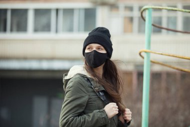 Kulak ilmiği maskeli, siyah şapkalı, arka planda bina olan genç bir kızın portresi. Siyah maskeli bir genç, dışarıda.