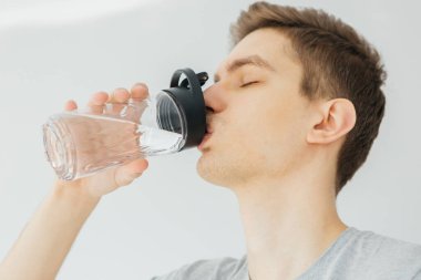 Spor sırasında gri gömlekli genç adam su içiyor. Evde spor var. Gri arka plandaki adam