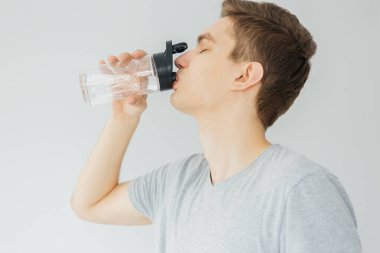 Spor sırasında gri gömlekli genç adam su içiyor. Evde spor var. Gri arka plandaki adam