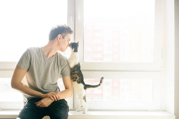 Genç adam pencere eşiğinde bir kediyle oturuyor. En sevdiği hayvan. Arka planda kedisi olan bir adam, güneş ışığı olan bir pencerenin arkasında.