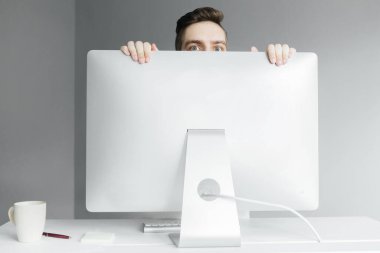 Gri arka planda bilgisayarın arkasından röntgencilik yapan genç bir adam. Masaüstünde çalışan bir işçi