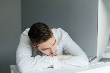 Ofisinde bilgisayarla beyaz gömlekli genç bir adam masaüstünde yatıyor. Gri arka planda çalışan bir işçi. Dinlenen adam