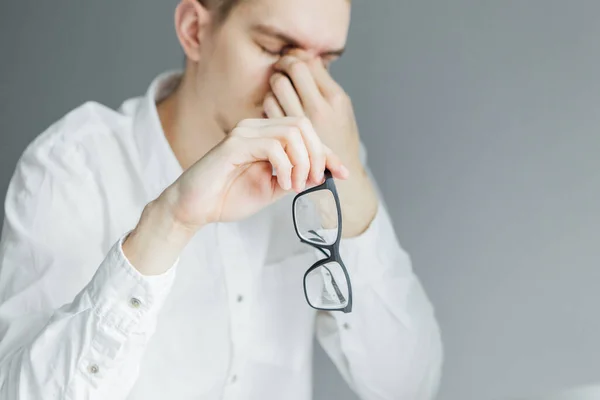 Genç adam göz yorgunluğu nedeniyle gözlüğünü çıkarıyor. Gri arkaplanda beyaz gömlekli işçi