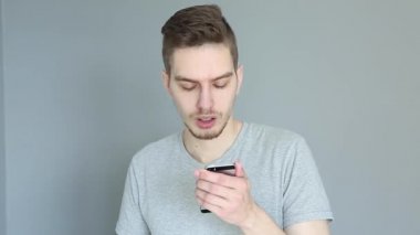 Gri arka planda telefonu olan, elini tutan şok olmuş genç bir adam. Ellerini başının üstüne koy. Gri gömlekli adam.