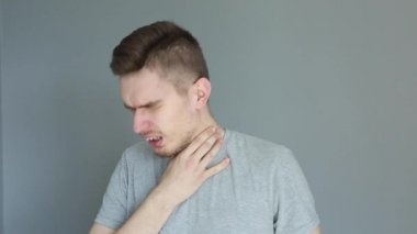 Gri arka planda gri gömlekli genç hasta adam öksürüyor ve kafasını tutuyor.