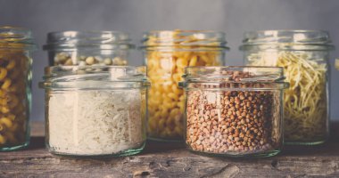 Tahta bir masada, cam kavanozlarda sağlıklı yiyecekler pişirmek için çeşitli çiğ tahıllar, taneler, fasulyeler ve makarna. Gri arka planda, temiz yemek, vegan, dengeli diyet, sıfır atık, çevre dostu, plastiksiz.