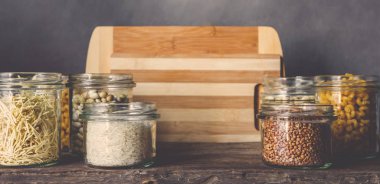 Tahta bir masada, cam kavanozlarda sağlıklı yiyecekler pişirmek için çeşitli çiğ tahıllar, taneler, fasulyeler ve makarna. Gri arka planda, temiz yemek, vegan, dengeli diyet, sıfır atık, çevre dostu, plastiksiz.