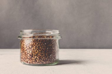 Tahta bir masada, cam kavanozlarda sağlıklı yiyecekler pişirmek için çeşitli çiğ tahıllar, taneler, fasulyeler ve makarna. Gri arka planda, temiz yemek, vegan, dengeli diyet, sıfır atık, çevre dostu, plastiksiz.