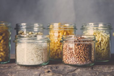 Tahta bir masada, cam kavanozlarda sağlıklı yiyecekler pişirmek için çeşitli çiğ tahıllar, taneler, fasulyeler ve makarna. Gri arka planda, temiz yemek, vegan, dengeli diyet, sıfır atık, çevre dostu, plastiksiz.