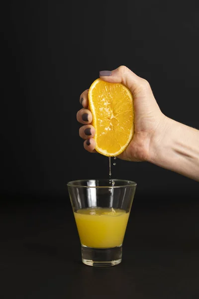 hand of a young woman squeezes juice from an orange into a glass, black background, vertical frame, closeup