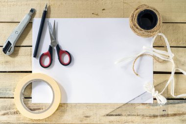 Merkezde beyaz bir kağıt tabakası var. Kenarlarında, ahşap arka planda bir el işi hediye kutusu yapmak için aletler var. Üst görünümde, boşluğu kopyalamak için.