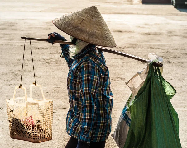 Stock image of unrecognizable Vietnamese with conical hat carries a yoke on her shoulder along the street. Profile wiew of a human with conical hat in contrast sunset light.