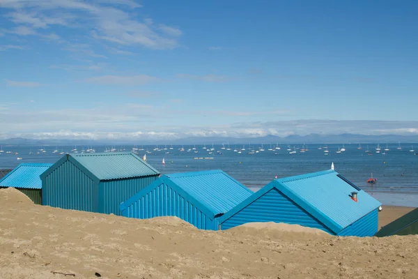 Blue beach huts in the sand look out over the sea with yachts and mountains in the distance at Abersoch, Gwynedd, Wales, UK.