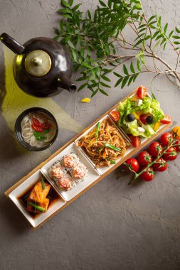 Tam öğle yemeği. Yunan salatası, cam erişte, rulo, somon filetosu, erişte çorbası ve gri zemin üzerinde çay olan çaydanlık..